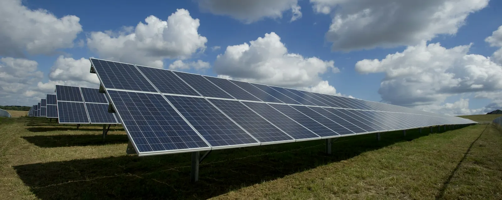 Rows of ground-mounted solar panels in a grassy field under a cloudy blue sky